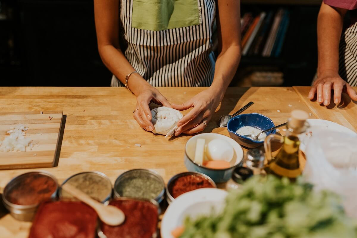 Woman forming dough in a cooking class setup with colorful spices, herbs, and ingredients on a wooden counter.